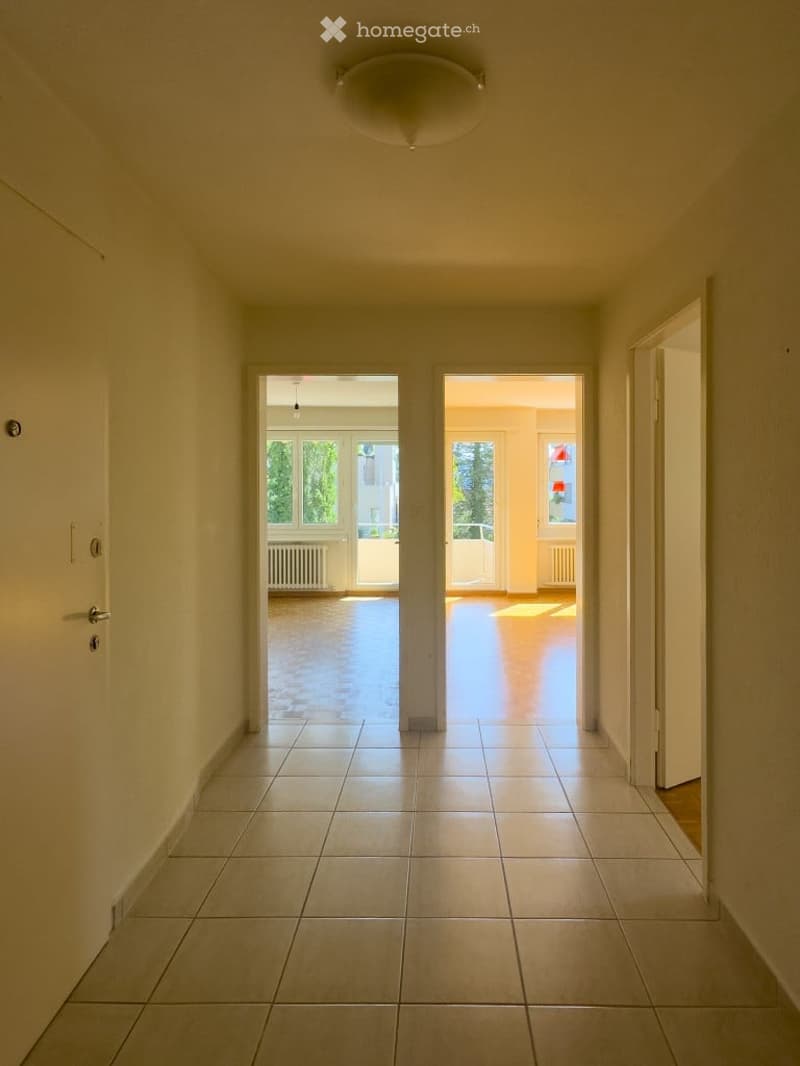 Empty hallway with tiled floor, white walls, and a ceiling light fixture. Two open doors lead to adjacent rooms with visible windows and radiators.