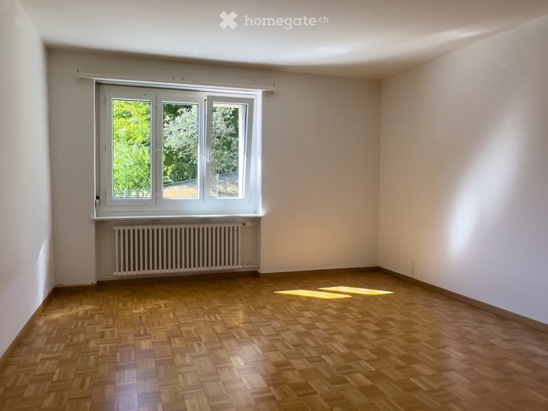 Empty room with wooden parquet flooring, white walls, and a large window with white frames. A radiator is visible below the window.