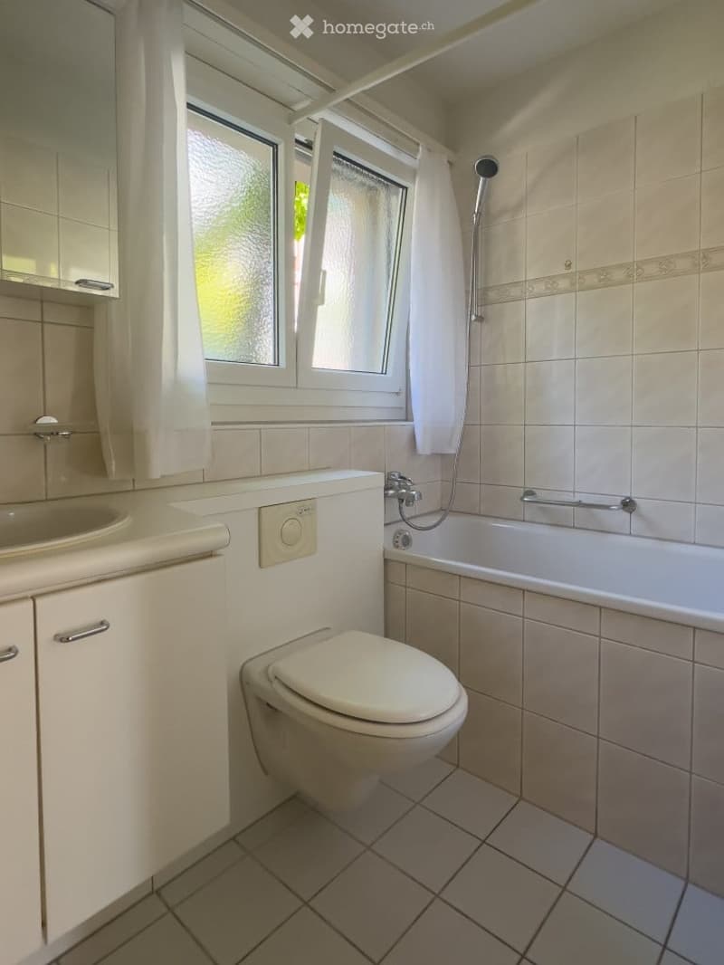 Bathroom with a toilet, bathtub, sink, mirror, and cabinet. The room features tiled walls and floor, a window with white curtains, and a showerhead above the bathtub.