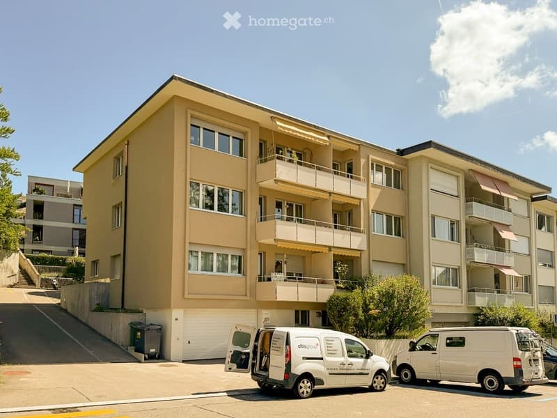 Multi-story residential building with beige facade, multiple windows, balconies, and a parking area with vans.