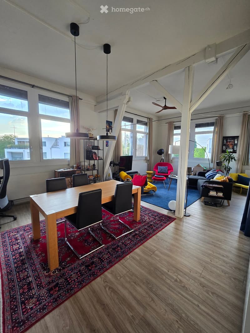 Open-plan living and dining area with wooden flooring, large windows, a dining table with chairs, a sofa, a red accent chair, a small table, and decorative rugs.