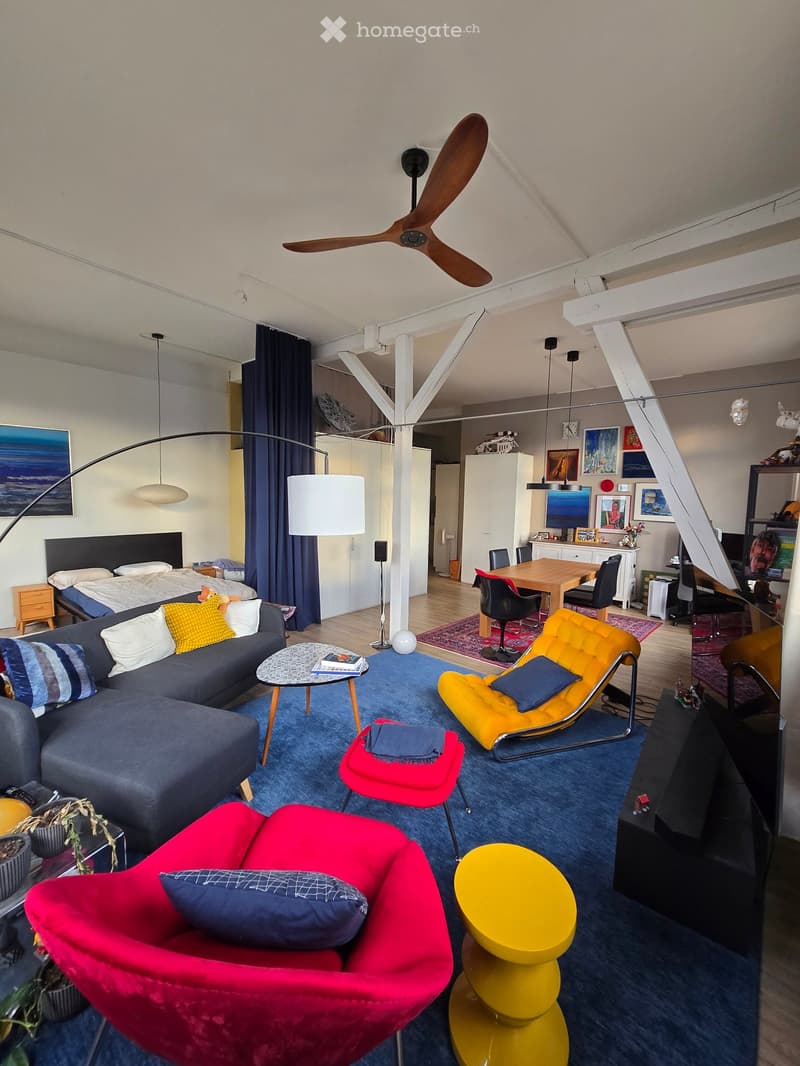 A modern, open-concept living and bedroom area with a grey sectional sofa, colorful chairs, a coffee table, a yellow accent chair, a red ottoman, a yellow round table, a ceiling fan, a bed with pillows, a nightstand, and a dining area with a table and cha