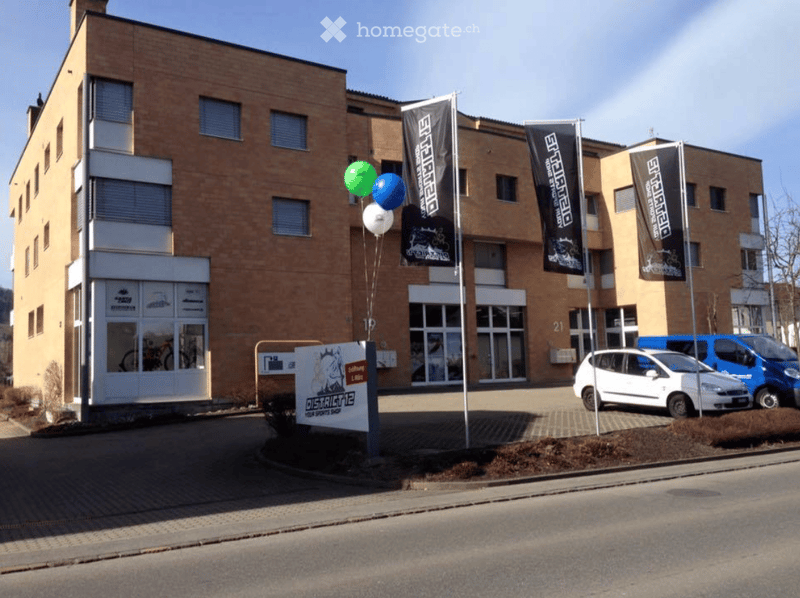 Modern multi-story building with brick facade, large windows, and signage for "District 12". Banners and balloons are displayed outside, with a small parking area containing cars.