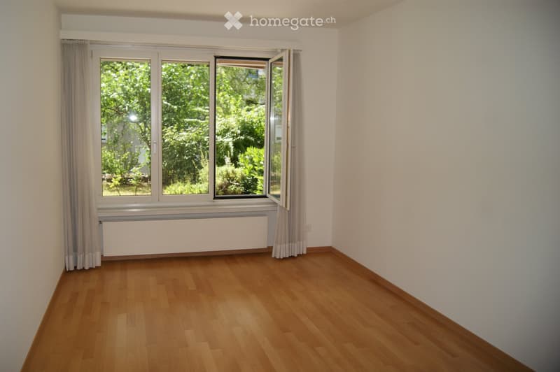 Empty room with wooden flooring, white walls, large window with curtains, radiator, and visible greenery outside.