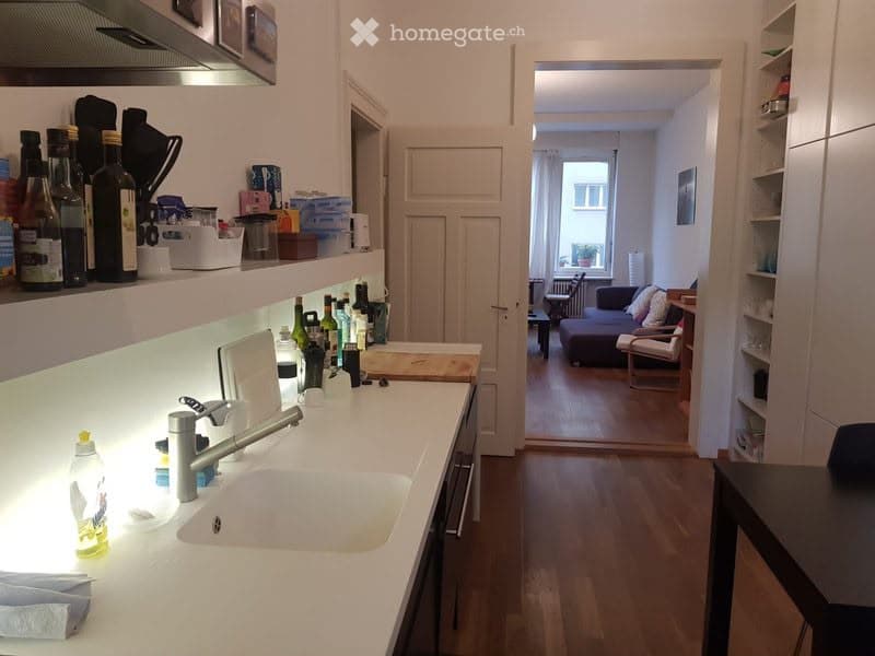 The image shows a modern, well-lit kitchen with white countertops, a sink, and various bottles and items on the shelves. An open doorway leads to a living area with a sofa, a chair, and a small table, all set on a wooden floor.