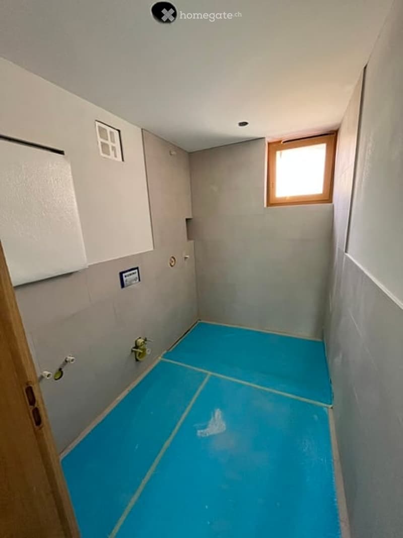 Empty room with light grey walls, blue tiled floor, small window, and a door.