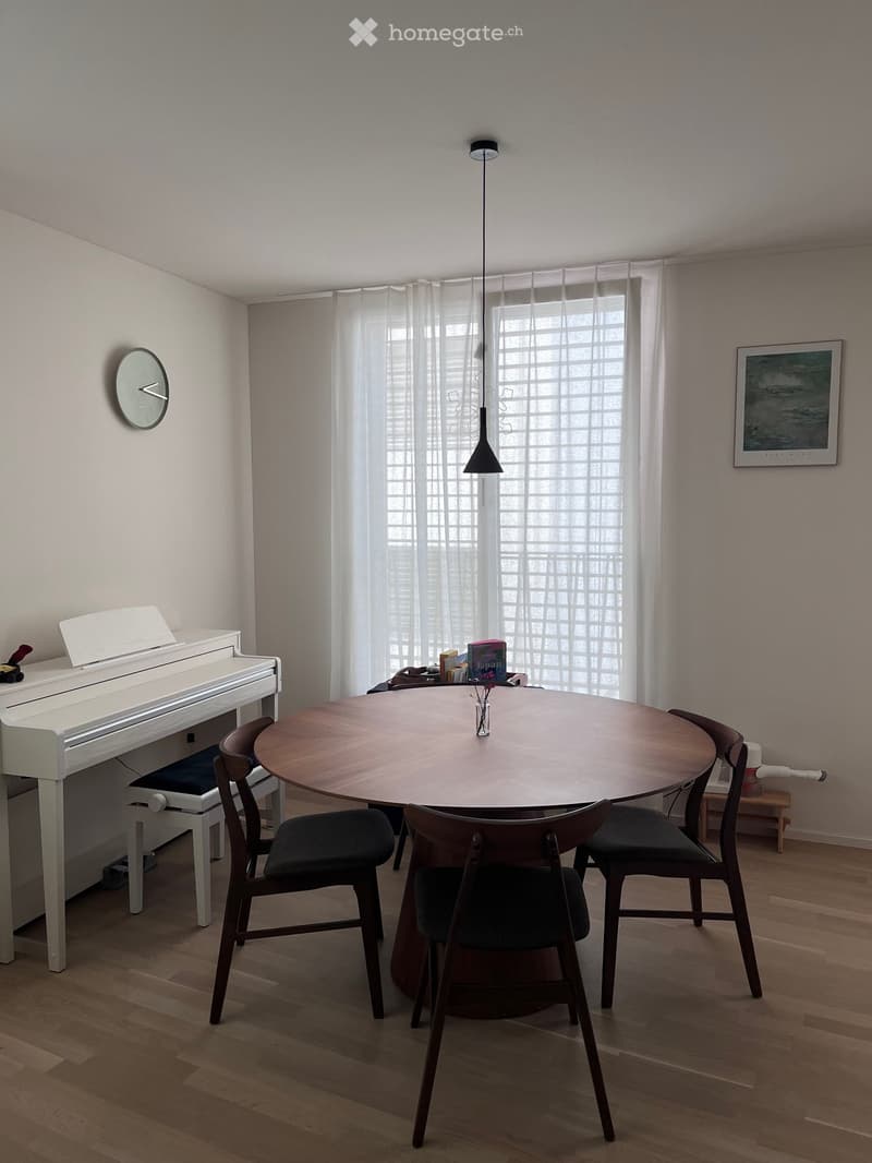 A modern dining area with a round wooden table surrounded by four chairs, a white piano, and a clock on the wall.
