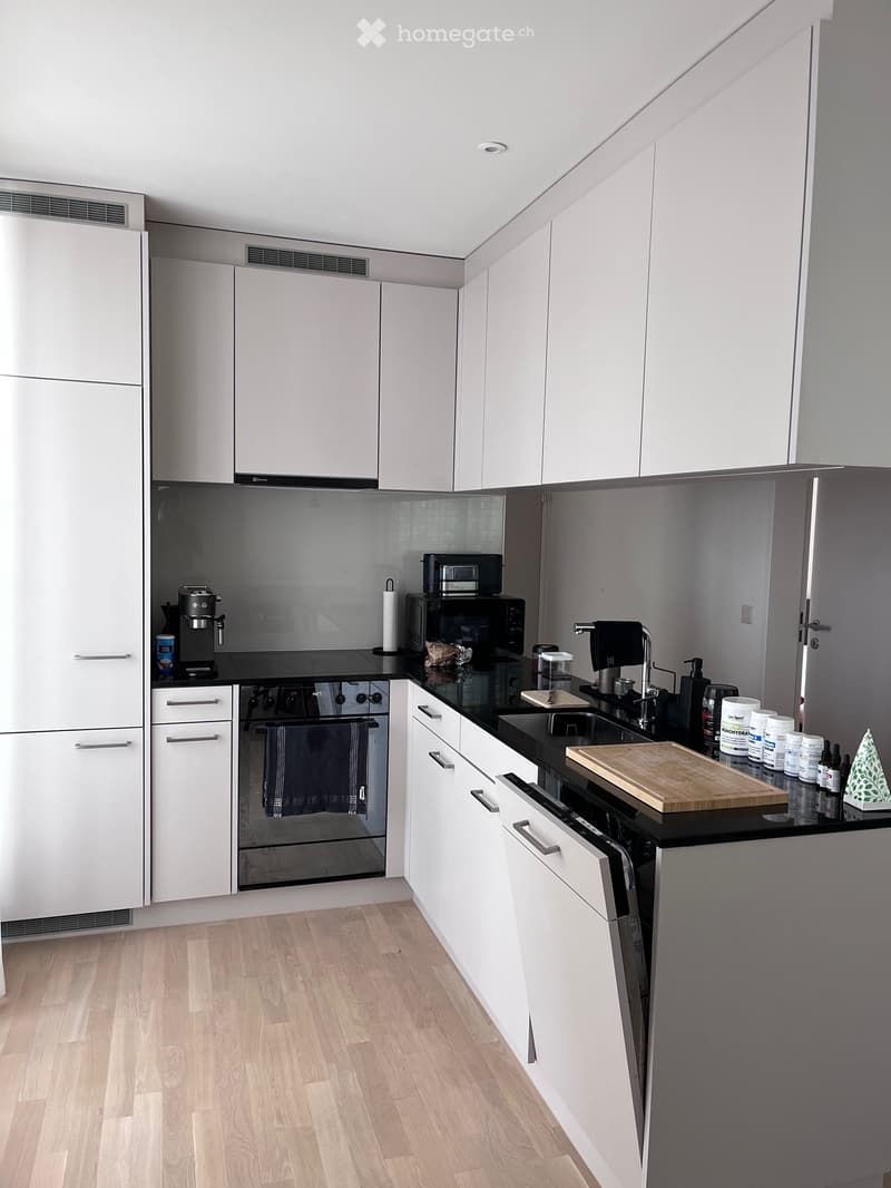 Modern kitchen with white cabinets, black countertops, and various appliances including a refrigerator, oven, microwave, and coffee maker. Wooden flooring and a sink with a faucet are also visible.