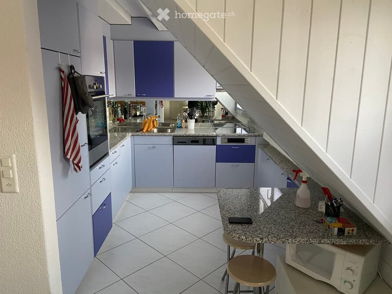 Modern kitchen with white and blue cabinets, granite countertops, and stainless steel appliances including an oven, dishwasher, and microwave. There are two stools at the island, a bottle, a cup, and a cellphone on the counter.