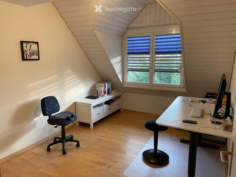 Bright, slanted-ceiling room with wooden flooring, a desk with a computer setup, a chair, a stool, a small cabinet, and a picture frame on the wall.