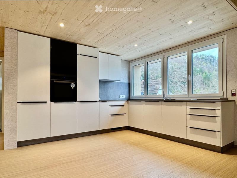 Modern kitchen with light wood cabinets, stainless steel appliances, and large windows overlooking a wooded area.