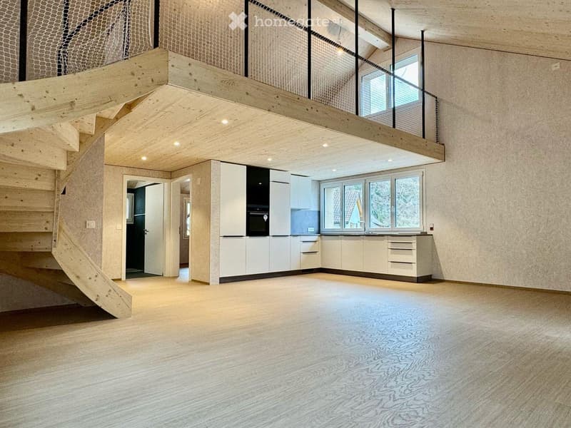 Open-plan living space with a modern kitchen area featuring white cabinets, an oven, and a sink, connected to a wooden staircase leading to an upper level with a balcony.