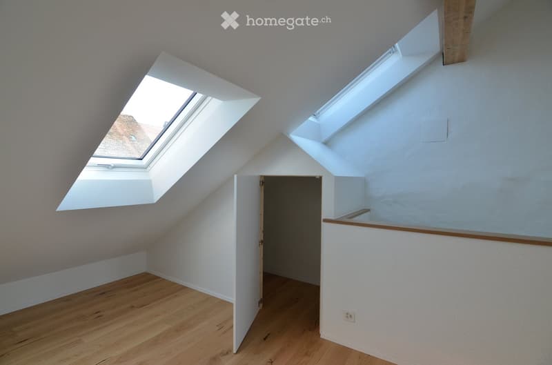 Empty attic room with wooden flooring, white walls, and two skylight windows. The room features exposed wooden beams and an open doorway leading to another space.