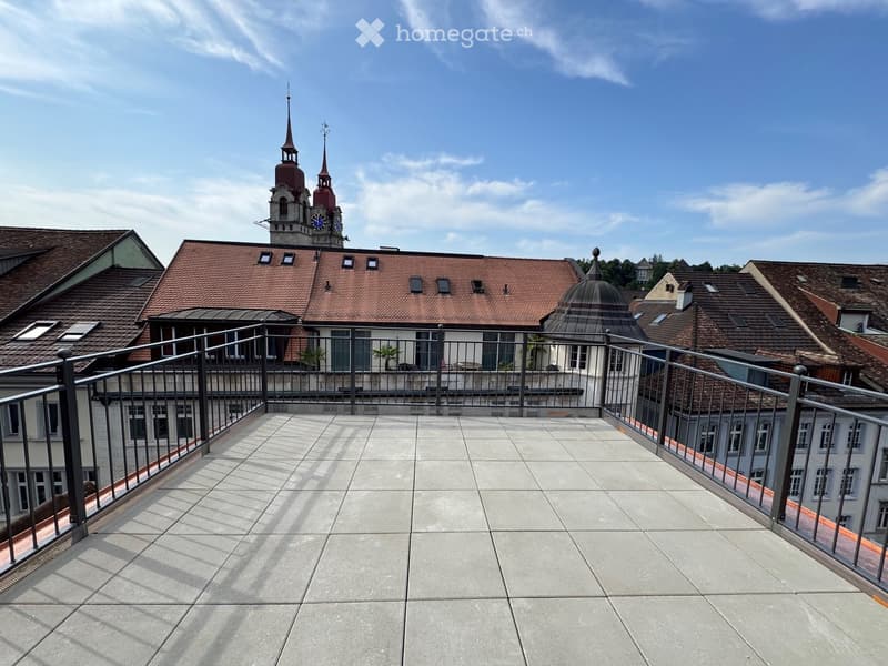 A spacious balcony with a modern metal railing, tiled floor, and a view of a historic building with a clock tower and surrounding rooftops.