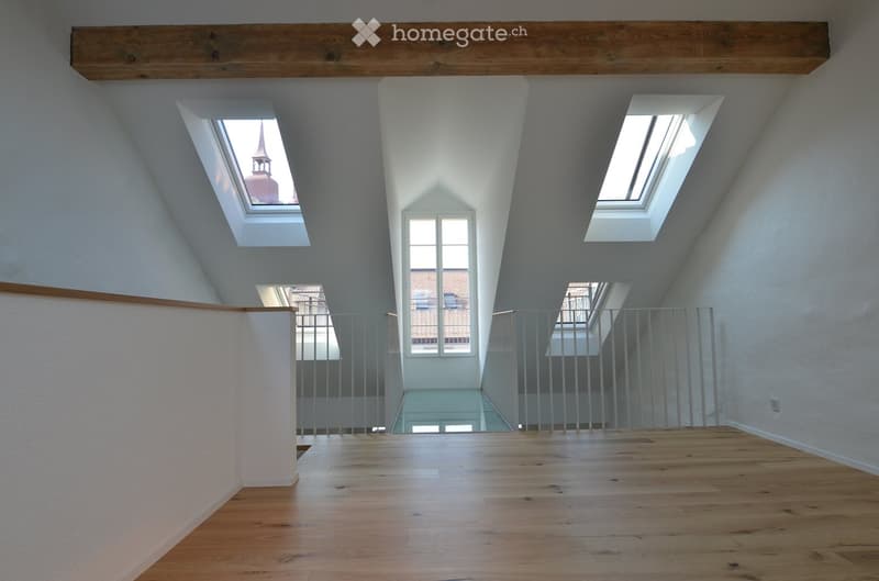 Bright, open loft space with wooden flooring, exposed wooden beams, and multiple skylights. The room features a modern metal railing and a view of the cityscape through the windows.