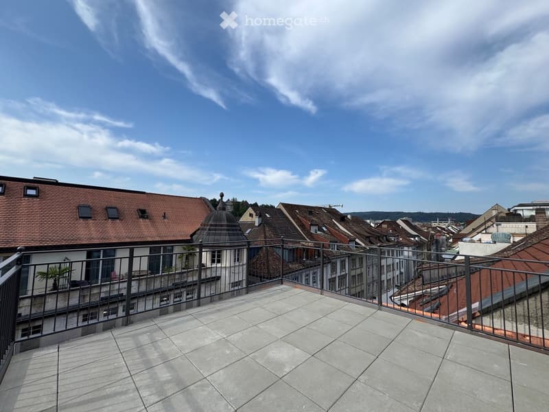 Balcony with tiled floor and metal railing, overlooking a cityscape with red-tiled roofs and a dome-shaped building.