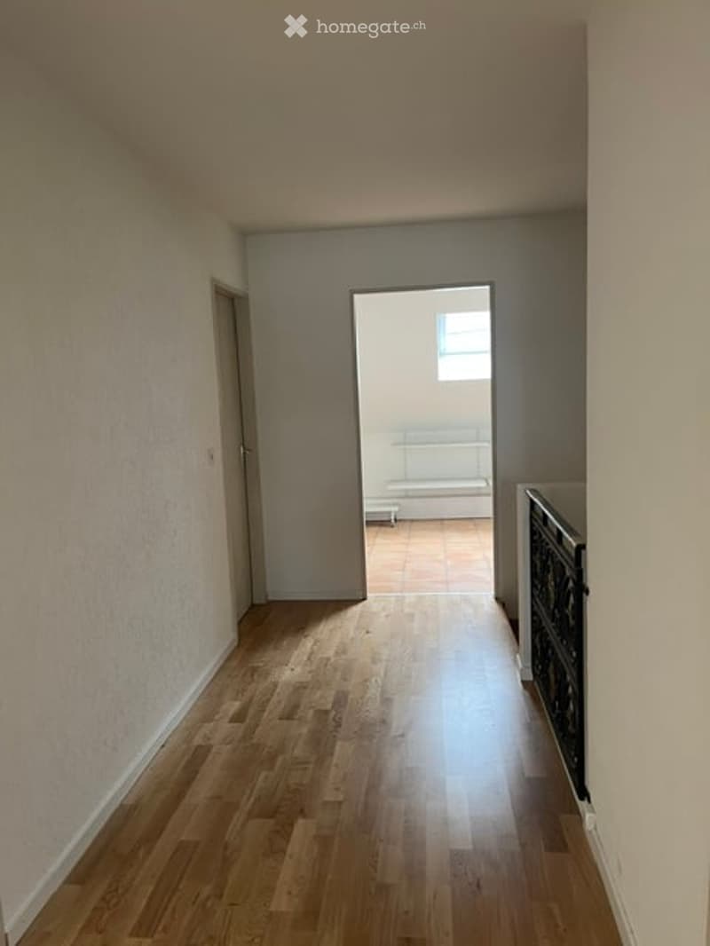 Empty hallway with wooden flooring and white walls, leading to an adjacent unfurnished room with a window and radiator.
