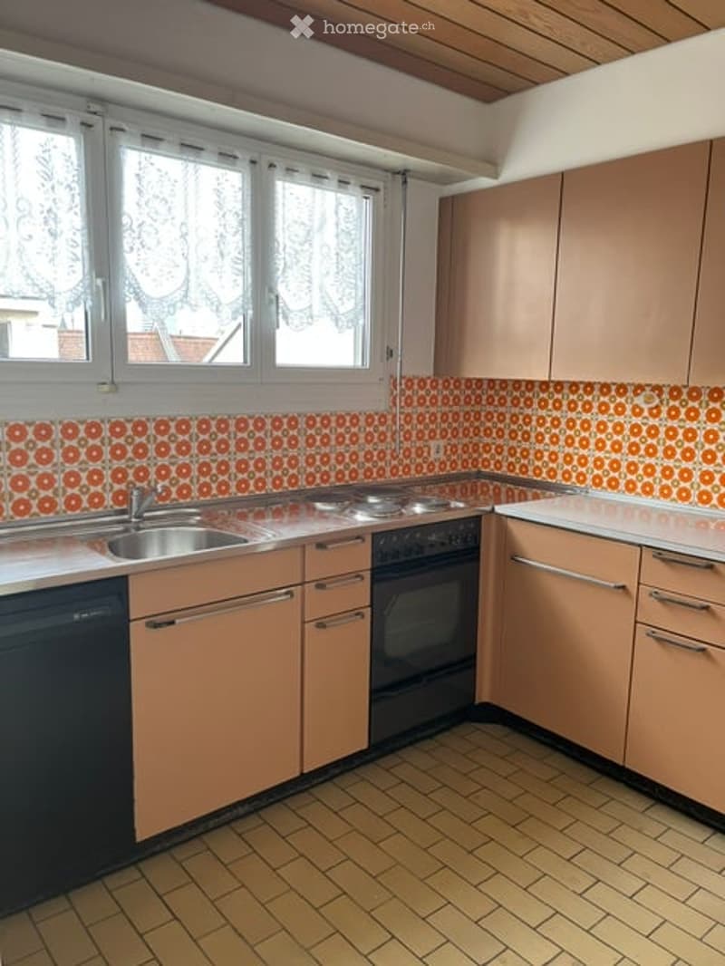 A compact kitchen with light pink cabinets, a white countertop, and orange patterned backsplash. Features include a sink, stove, oven, and dishwasher.