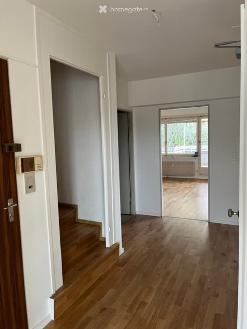 Empty hallway with wooden flooring, white walls, and a staircase leading upwards. A doorway leads to another room with a visible radiator and window.