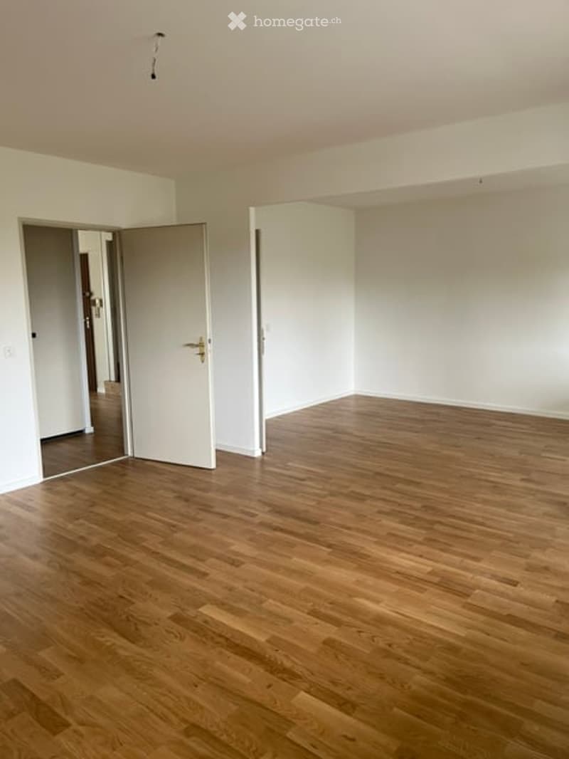 Empty room with wooden flooring and white walls, featuring an open doorway leading to another room.