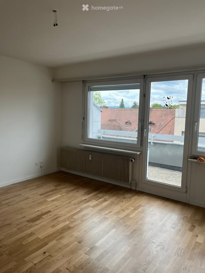 Empty room with wooden flooring, white walls, large window with view, radiator, and sliding glass door leading to balcony.
