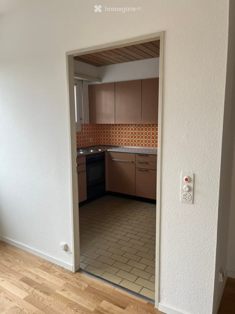 A small kitchen with light brown cabinets, a tiled backsplash, and a wooden floor. An oven is visible, and the space is connected to a hallway with wooden flooring.