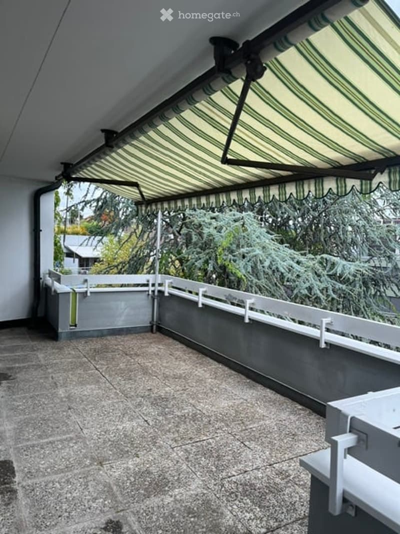 Balcony with striped awning, tiled floor, white railing, and view of greenery.