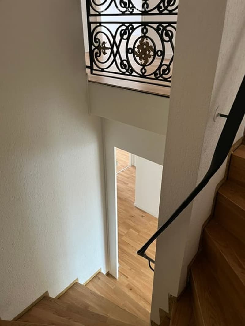 Spiral staircase with wooden steps and a black wrought-iron railing, leading to an open doorway with light-colored walls and wooden flooring visible beyond.
