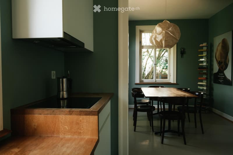 A modern kitchen and dining area with dark green walls, a wooden countertop, a pendant light, and a dining table with chairs.