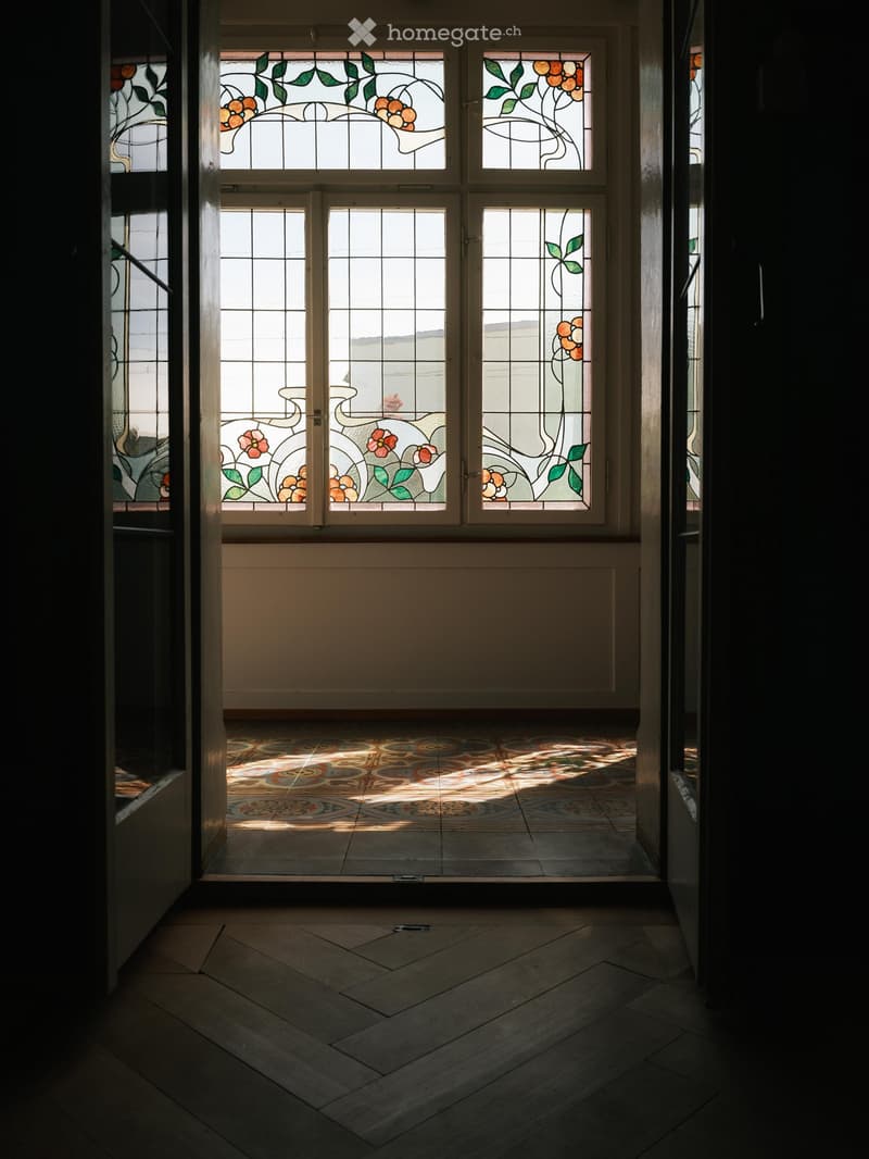 Hallway with stained glass windows, patterned tile floor, and wooden flooring leading to another area.