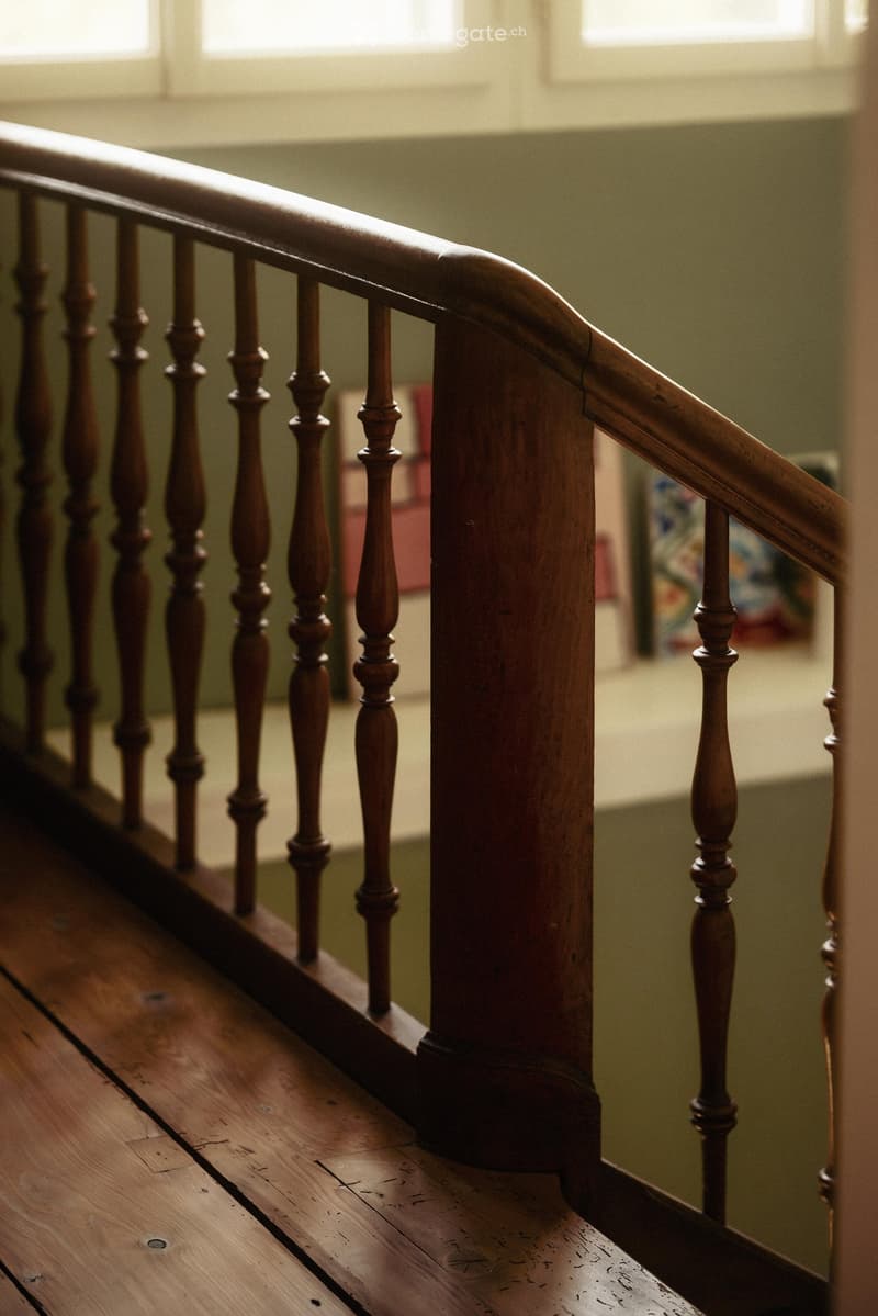 Wooden staircase with a polished handrail and balusters, leading upwards with natural light from windows above.