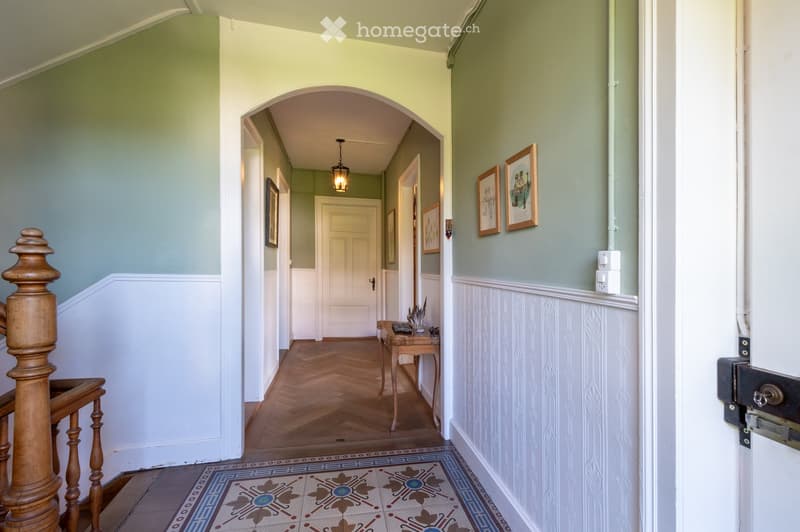 Elegant hallway with wooden herringbone flooring, decorative patterned tiles, framed artwork, and a small wooden table with items on it.