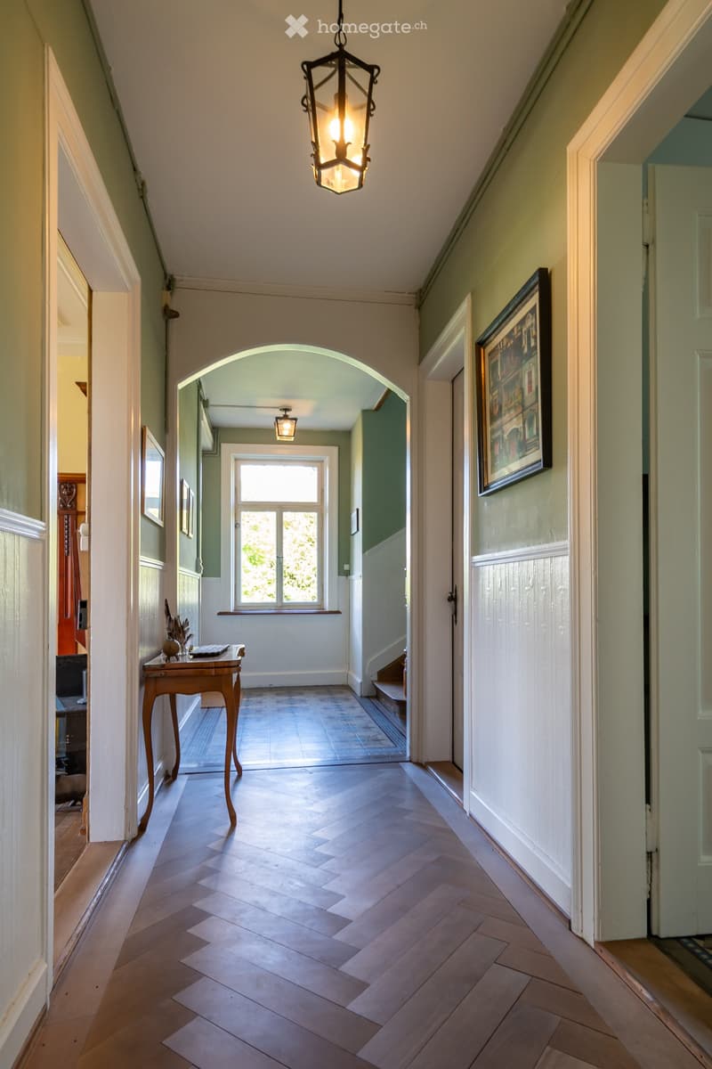 Elegant hallway with herringbone wooden flooring, arched doorway leading to another room, framed artwork on the wall, small wooden table with decorative items, and a hanging lantern-style light fixture.