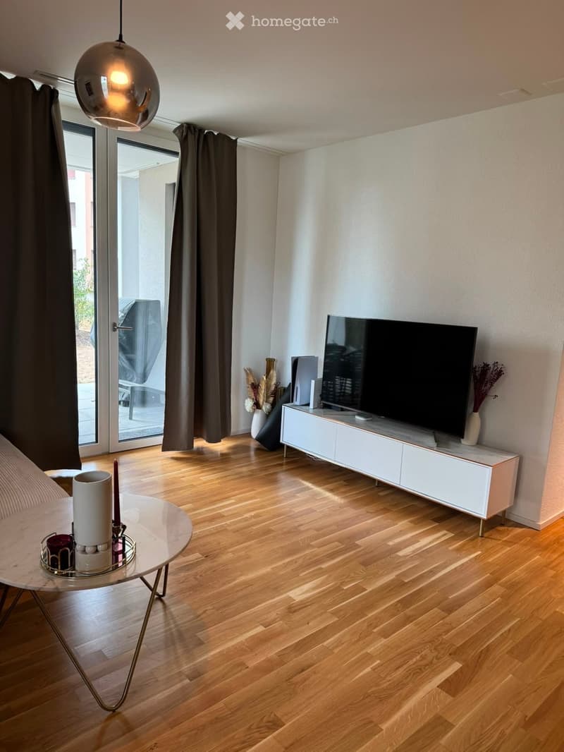 Modern living room with wooden flooring, large window with dark curtains, white TV cabinet, round glass coffee table, and decorative items.