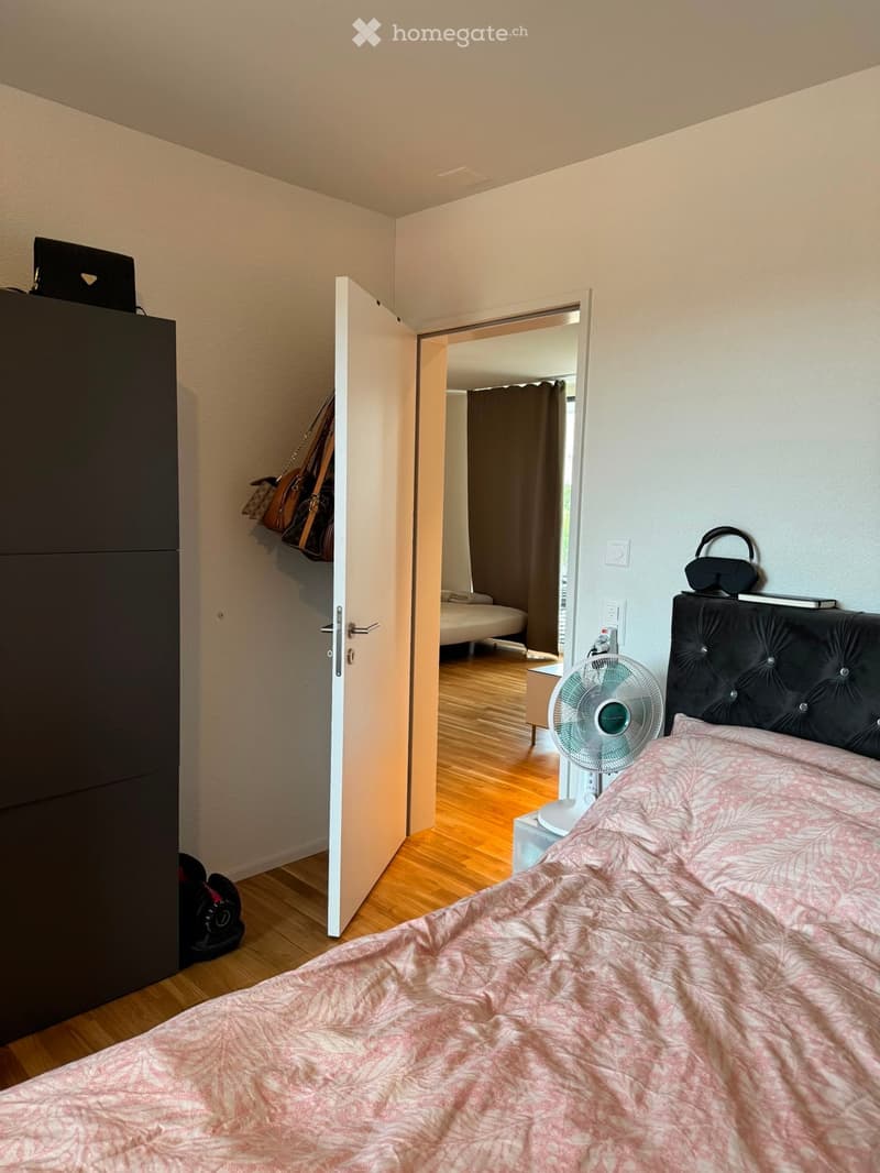 A modern bedroom with a wooden floor, a large bed with a pink patterned duvet, a black headboard, a standing fan, a wardrobe, and a laptop on top. An open door leads to another room with a bed and a curtain.