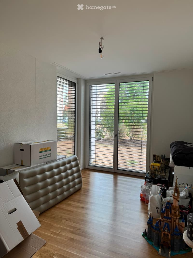Bright living room with wooden flooring, large windows, and a sliding glass door leading to a balcony. Visible items include a cream-colored sofa, a small table with toys, and several storage boxes.