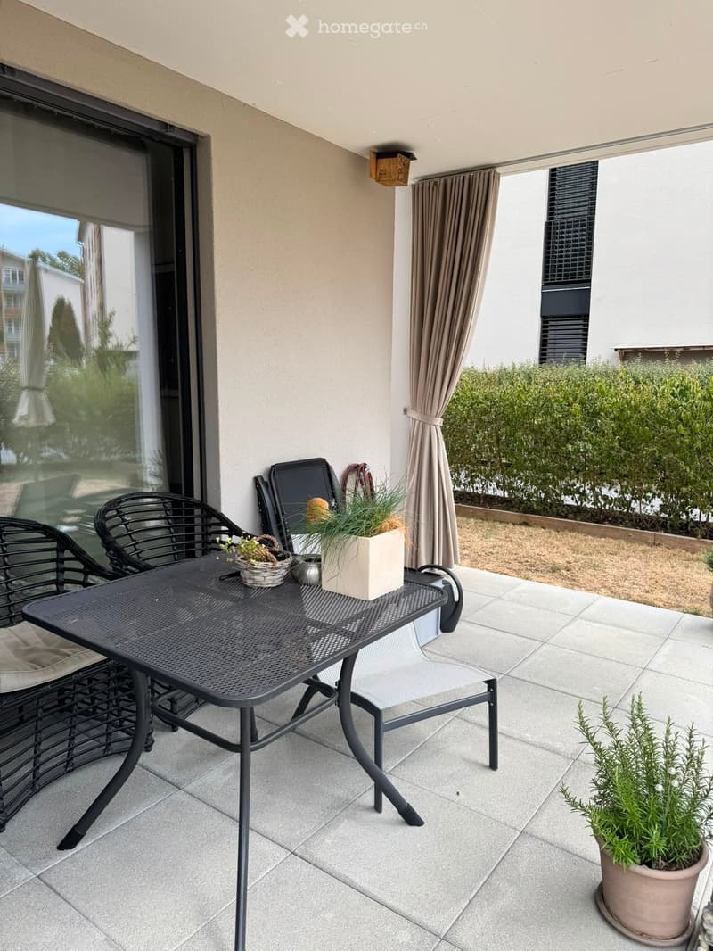 Balcony with a black metal table, two chairs, and potted plants, with a view of greenery outside.