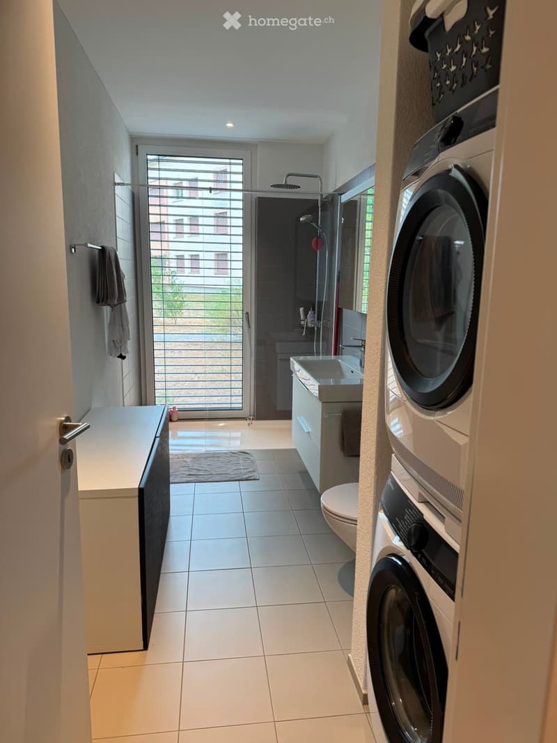 Modern bathroom with white tiles, a sink, toilet, shower, washing machine, and a towel hanging on the wall.