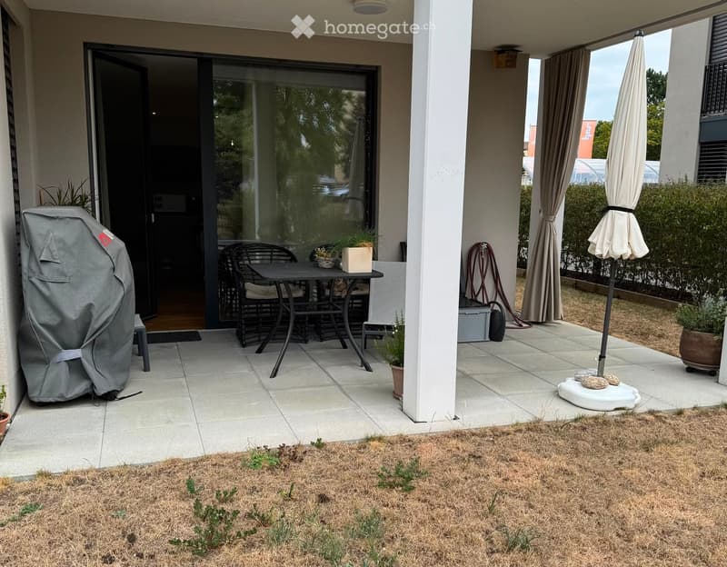 Balcony with a table, chairs, umbrella, and a covered grill. There is a view of the garden and surrounding area.