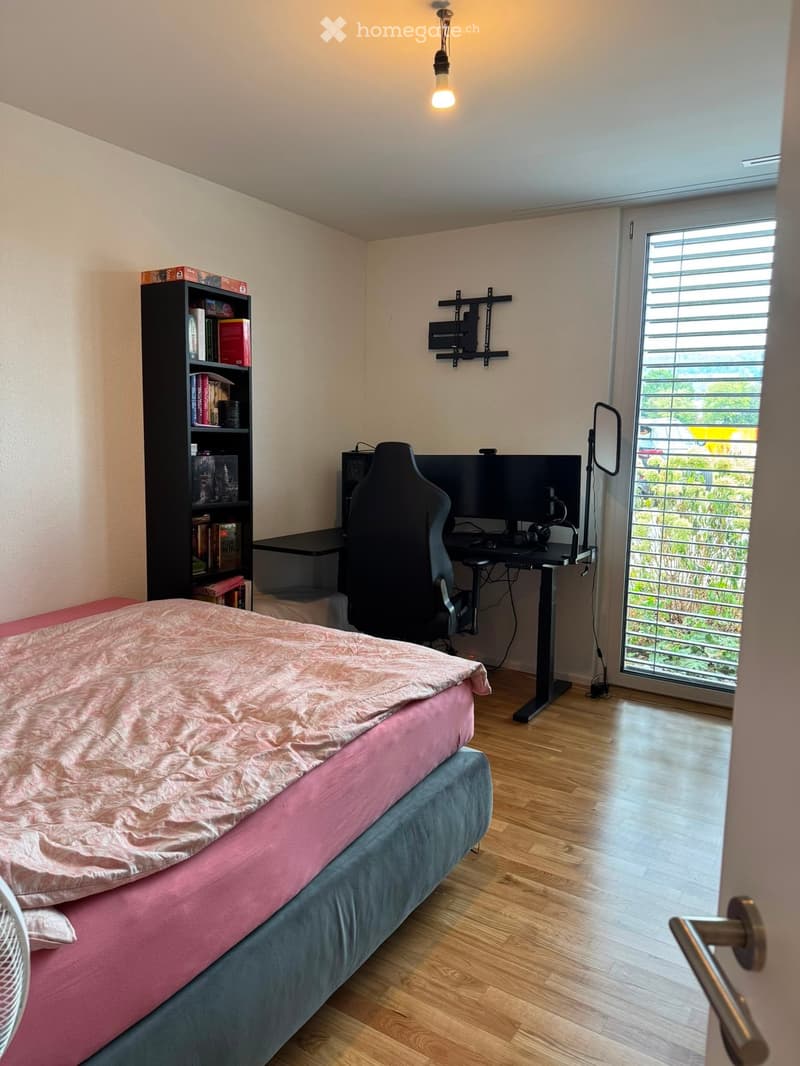 A modern bedroom with a bed, desk, chair, bookshelf, and a large window with blinds. The room features wooden flooring and a hanging light fixture.