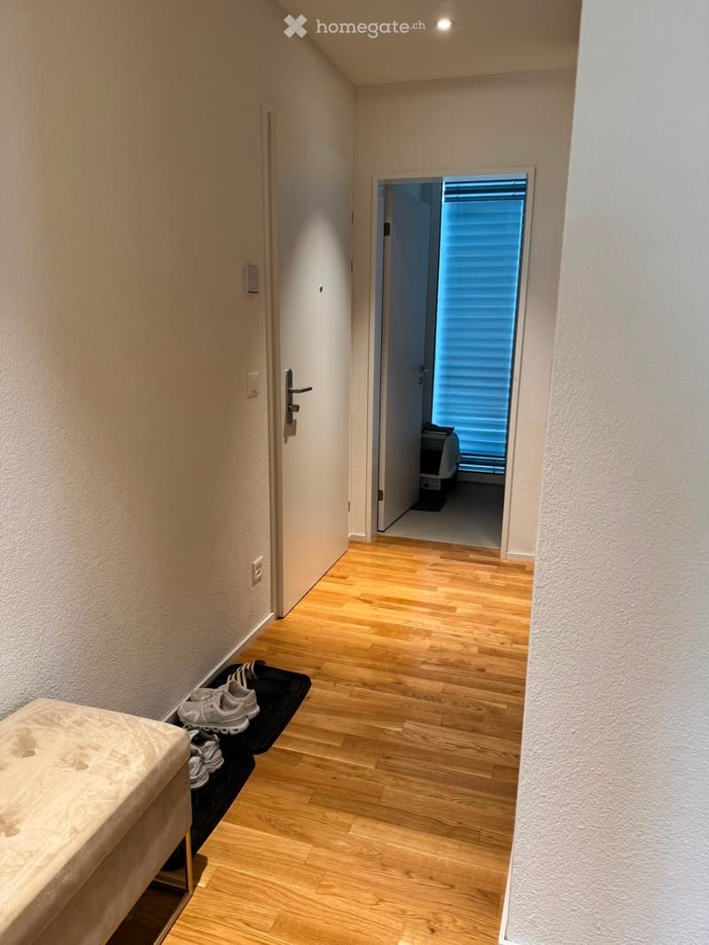 A narrow hallway with wooden flooring, white walls, and a small bench with shoes placed on it. A door leads to another room with blinds covering the window.