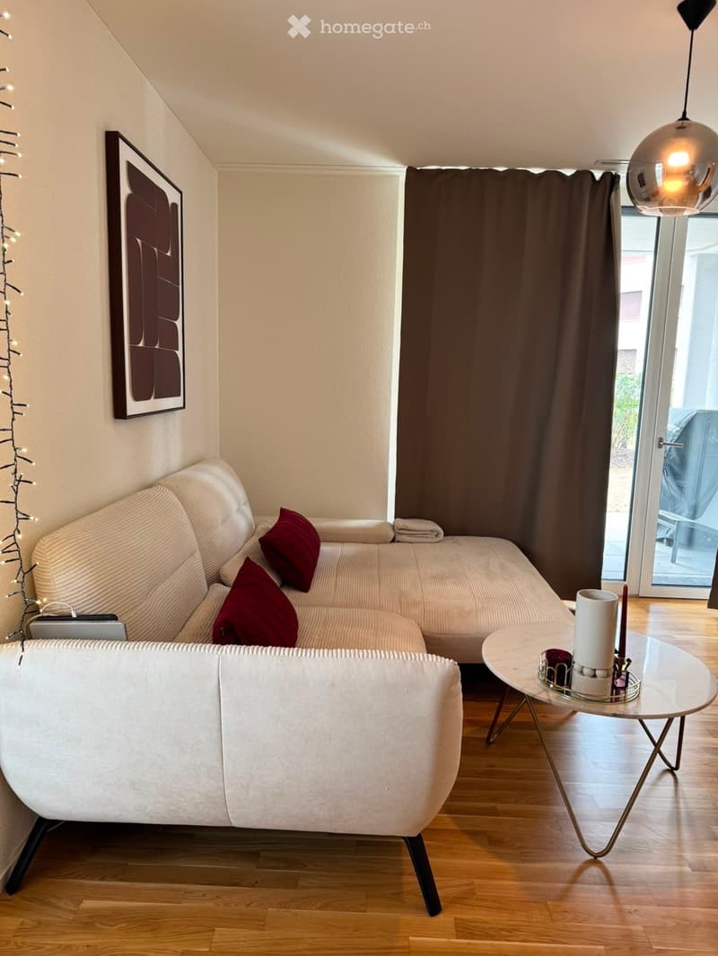 Modern living room with a white sectional sofa, red throw pillows, a round glass coffee table with candles, a picture frame on the wall, and a laptop on the floor. The room has wooden flooring and large windows with dark curtains.