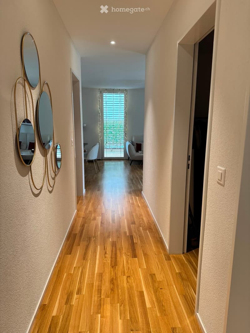 A modern hallway with light wooden flooring, beige walls, and a decorative wall-mounted mirror set. The hallway leads to a room with a white chair and a small table visible at the end.