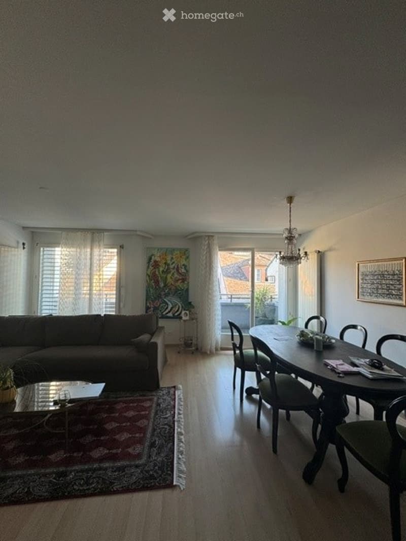 Open-plan living and dining area with a dark brown sectional sofa, coffee table, dining table with chairs, chandelier, and artwork on the walls.