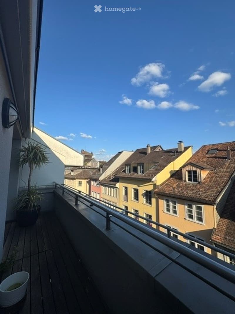 Balcony with wooden decking, metal railing, potted plants, overlooking a cityscape with colorful buildings and rooftops.