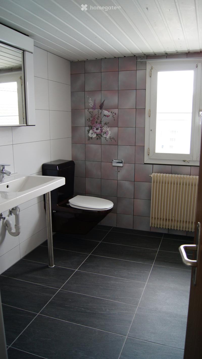 Modern bathroom with dark grey tiles, white sink, toilet, and radiator. Features a floral design on the wall tiles and a large window letting in natural light.