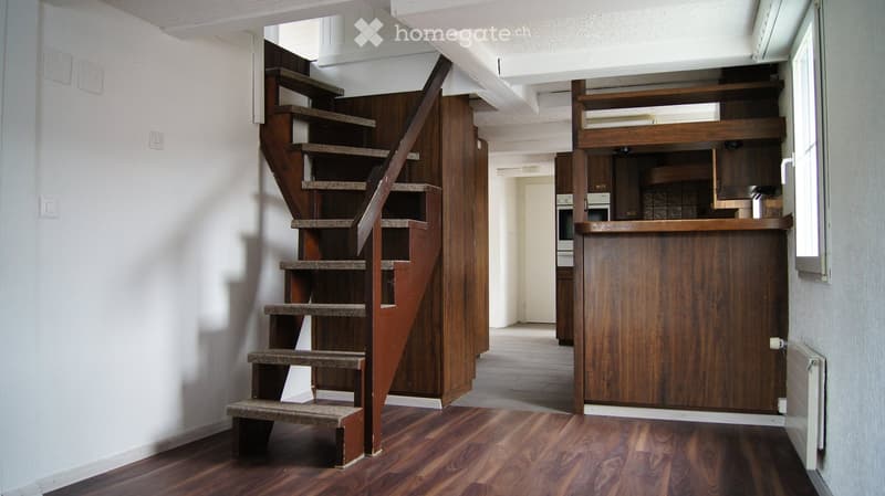 Spacious hallway with wooden flooring, a wooden staircase leading upwards, and a wooden cabinet with shelves and a countertop. The walls are white, and there is a visible doorway leading to another room.