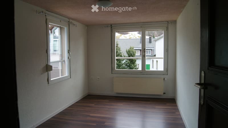 Empty room with wooden flooring, white walls, and large windows offering a view of neighboring buildings and greenery.
