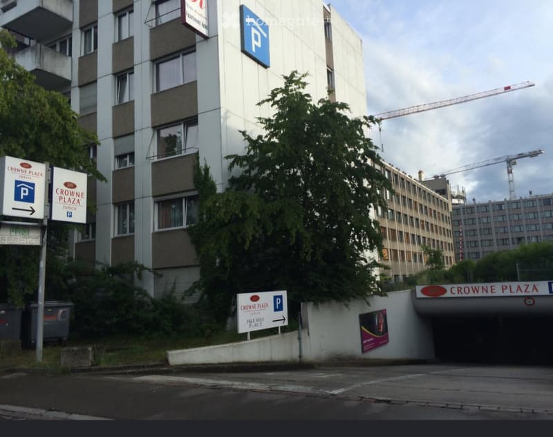 Multi-story building with signage for "Crowne Plaza Zurich" and parking directions, surrounded by greenery and a construction crane in the background.