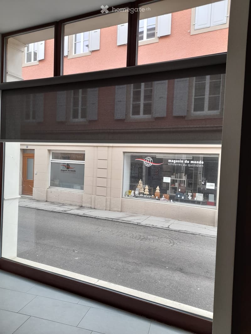 Balcony overlooking a street with a shop named "magasin du monde" visible in the reflection. The balcony has large glass panels and a wooden frame.